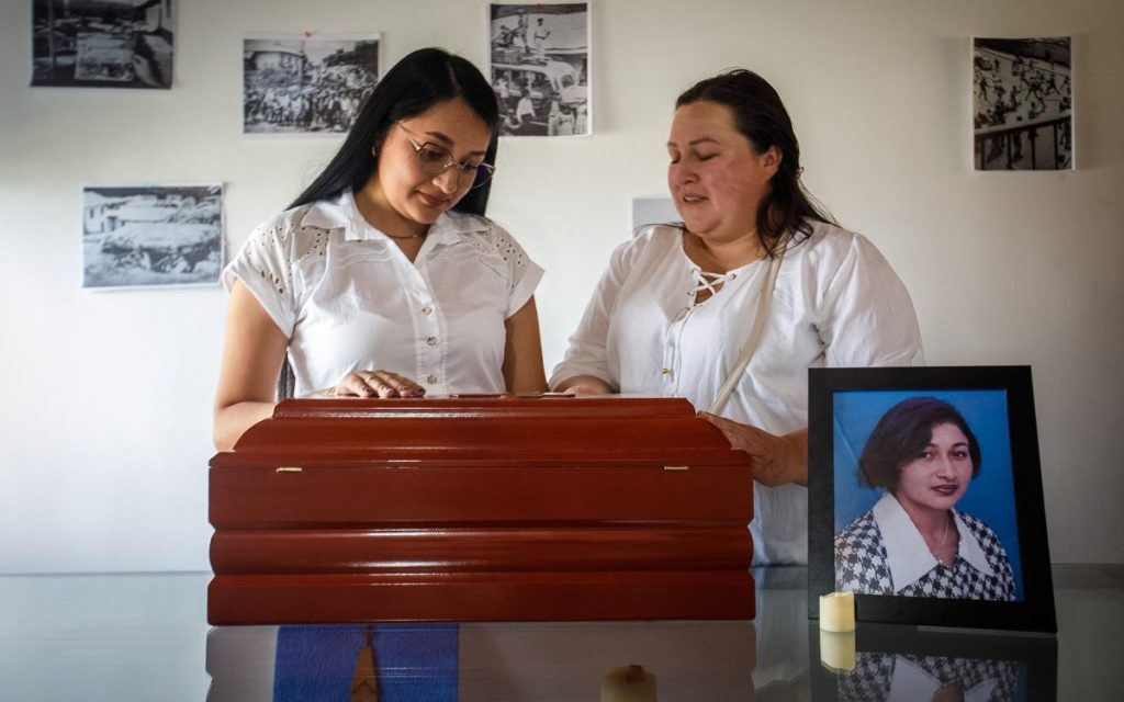 Dos mujeres vestidas de blanco miran con tristeza y respeto un pequeño cofre fúnebre de madera ubicado sobre una mesa de vidrio en un espacio interior. La mujer de la izquierda, con gafas y cabello largo y oscuro, apoya suavemente su mano sobre el cofre con la mirada baja. A su lado, la otra mujer la acompaña con un gesto de resignación y dolor. A la derecha de la mesa, en primer plano, se observa un portarretratos con la fotografía en color de una mujer de cabello corto y saco de cuadros, junto a una pequeña vela encendida. En la pared del fondo se aprecian varias fotografías pequeñas en blanco y negro que retratan momentos históricos o comunitarios.