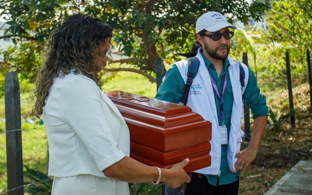 En un espacio exterior, una mujer de espaldas y perfil, vestida de blanco y con cabello rizado, sostiene con ambas manos un pequeño cofre de madera con una cruz tallada en la parte superior. Frente a ella, un hombre con gorra y chaleco blanco de la Unidad de Búsqueda de Personas dadas por Desaparecidas (UBPD), que lleva también una camisa verde, morral y carné de identificación al cuello, la observa con atención mientras apoya una mano en su cadera. Al fondo se observa vegetación, un árbol, postes y un cerco de alambre, bajo una luz natural de día.