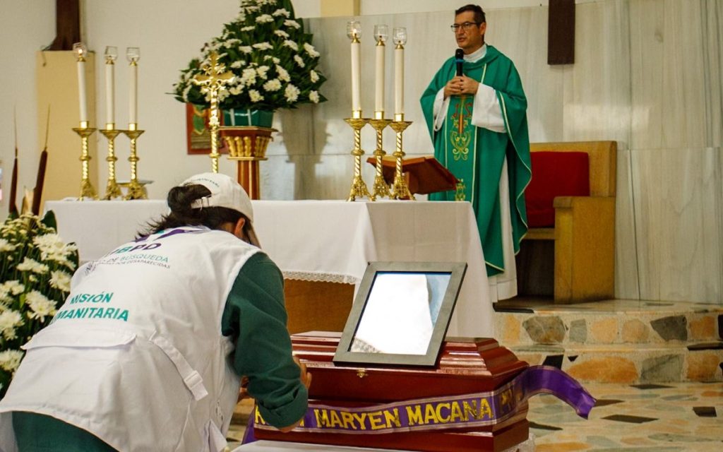 Perspectiva desde el interior de una iglesia durante una ceremonia. En primer plano y de espaldas, una persona con gorra y chaleco blanco de la "Misión Humanitaria" de la Unidad de Búsqueda de Personas dadas por Desaparecidas (UBPD) se encuentra agachada o inclinada frente al altar, manipulando o decorando un pequeño cofre fúnebre de madera. El cofre tiene encima un portarretratos y está adornado con una cinta morada y dorada que lleva el nombre "MARYEN MACANA". Al fondo, sobre el presbiterio, un sacerdote con ornamentos verdes habla por micrófono frente al altar, que está decorado con grandes arreglos de flores blancas, candelabros dorados con velas encendidas y un crucifijo.