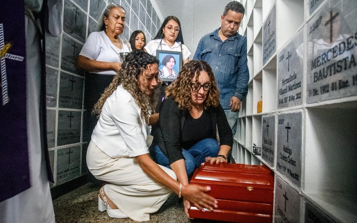 En el pasillo estrecho de un cementerio con nichos o tumbas numeradas en las paredes, dos mujeres se encuentran agachadas en el suelo, introduciendo o acomodando conmovidas un pequeño cofre fúnebre de madera en uno de los compartimentos inferiores. La mujer de la izquierda, con chaqueta blanca y pantalón claro, y la de la derecha, con gafas y ropa oscura, tocan con delicadeza el cofre con visibles muestras de dolor. Detrás de ellas, un grupo de familiares y acompañantes observa con tristeza, entre ellos una mujer que sostiene un portarretratos con la fotografía de una mujer, otra mujer mayor con una vela, una joven y un hombre con camisa de jean. A la izquierda se alcanza a ver el costado de un sacerdote con estola morada. En las lápidas de la derecha se leen nombres como 
