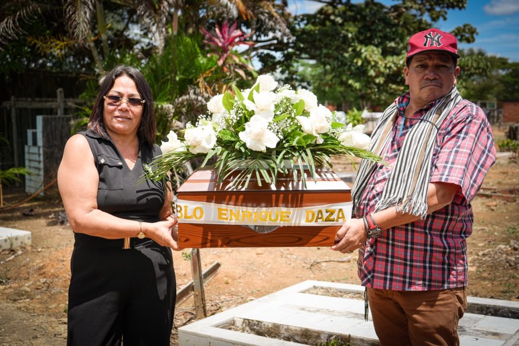 Fotografía en exteriores de una mujer a la izquierda, vestida de negro con gafas de sol, y un hombre a la derecha con camisa a cuadros y gorra roja, sosteniendo un pequeño cofre mortuorio de madera. Encima del cofre hay un arreglo de rosas blancas y una cinta decorativa que dice «Pablo Enrique Daza».