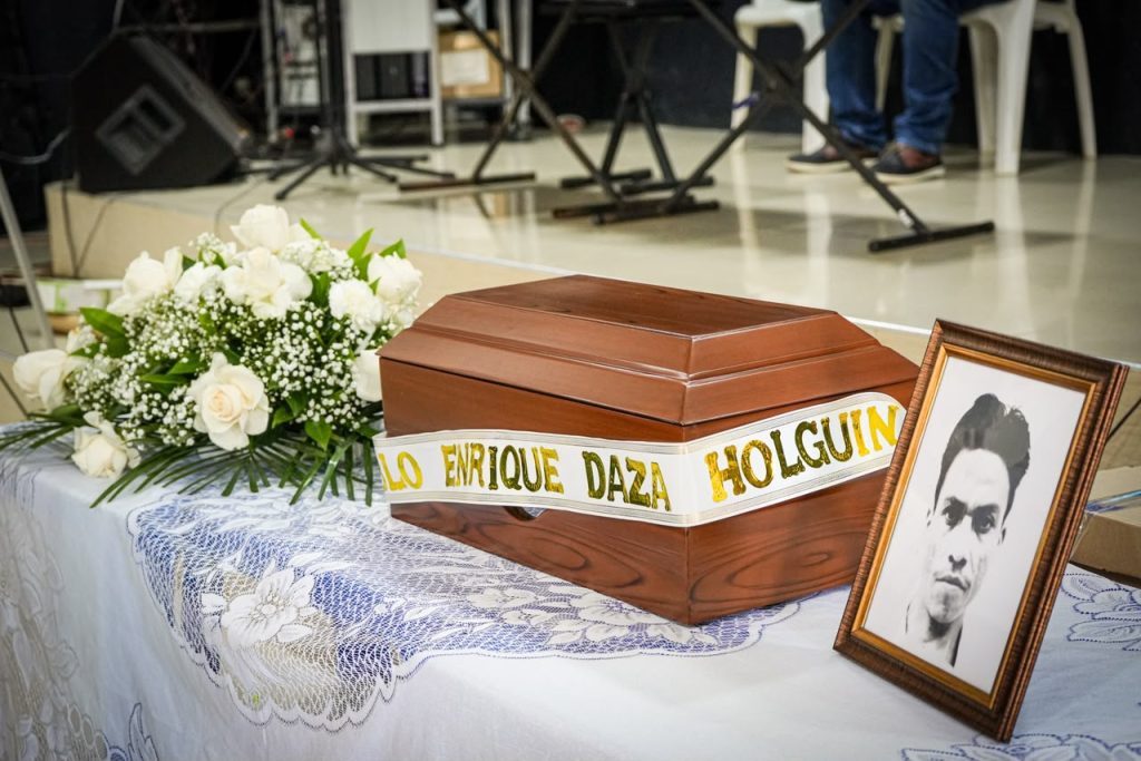 Toma en plano medio sobre una mesa con mantel blanco bordado. Se observa un cofre de madera con una cinta blanca de letras doradas que dice «Pablo Enrique Daza Holguín». A la derecha, un retrato enmarcado en blanco y negro de un hombre, y a la izquierda, un ramo de rosas blancas.