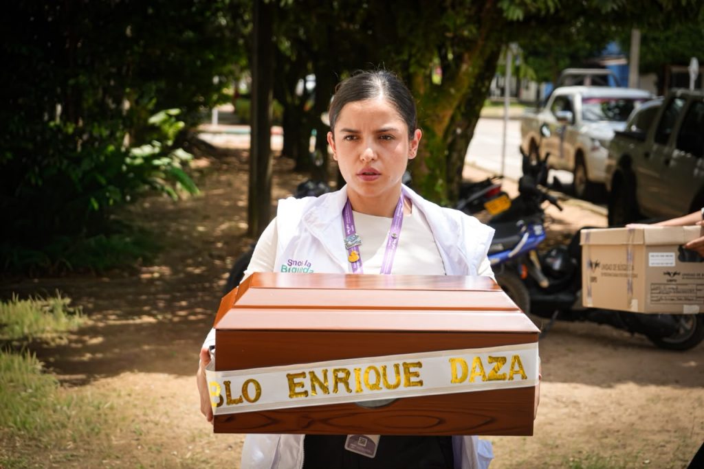 Una mujer joven que lleva puesto un chaleco blanco con el logo «Somos la Búsqueda» camina sosteniendo cuidadosamente frente a ella un cofre de madera para restos, el cual tiene una cinta con el nombre «Pablo Enrique Daza» impreso en letras doradas.