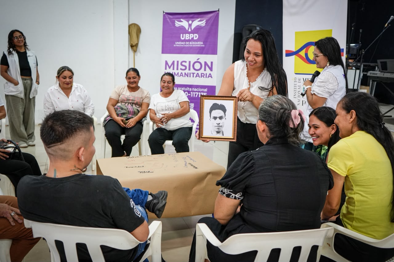 Reunión en un espacio interior con un pendón al fondo de la UBPD (Unidad de Búsqueda de Personas dadas por Desaparecidas). En el centro, una mujer sonriente de pie exhibe un retrato enmarcado ante un grupo de personas sentadas en círculo.