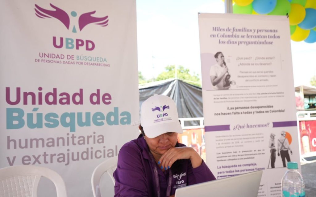 En el estand de la Unidad de Búsqueda de Personas dadas por Desaparecidas (UBPD), una mujer con gorra blanca y polo morado con el logo de la entidad mira atentamente la pantalla de su portátil. A sus lados hay pendones informativos de la UBPD con el lema «Unidad de Búsqueda humanitaria y extrajudicial» y preguntas dirigidas a familiares de personas desaparecidas en Colombia.
