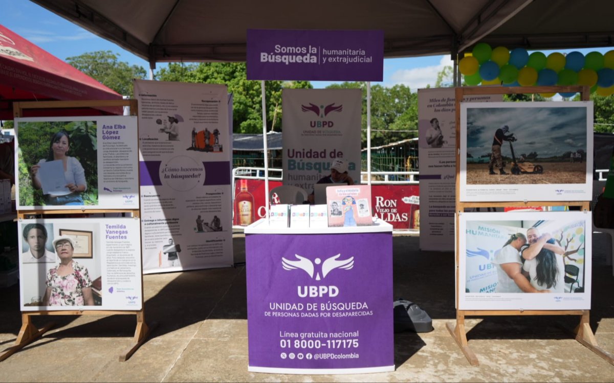Vista general del estand de la Unidad de Búsqueda de Personas dadas por Desaparecidas (UBPD). En el centro destaca un módulo morado con el logo institucional y el número de línea gratuita nacional 01 8000-117175. Alrededor hay carteleras con rostros e historias de personas buscadas, así como fotografías de labores de campo y reuniones comunitarias.