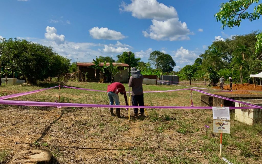 Vista panorámica de una zona rural a campo abierto bajo un cielo despejado. En el centro, dos personas instalan o ajustan un prisma y equipo topográfico en un terreno delimitado por una cinta de demarcación de color rosado. En la esquina inferior derecha hay un letrero técnico con el logotipo de la UBPD (Unidad de Búsqueda de Personas dadas por Desaparecidas) y el texto "AREA 01". Al fondo se observa vegetación frondosa, árboles y una pequeña edificación campesina.