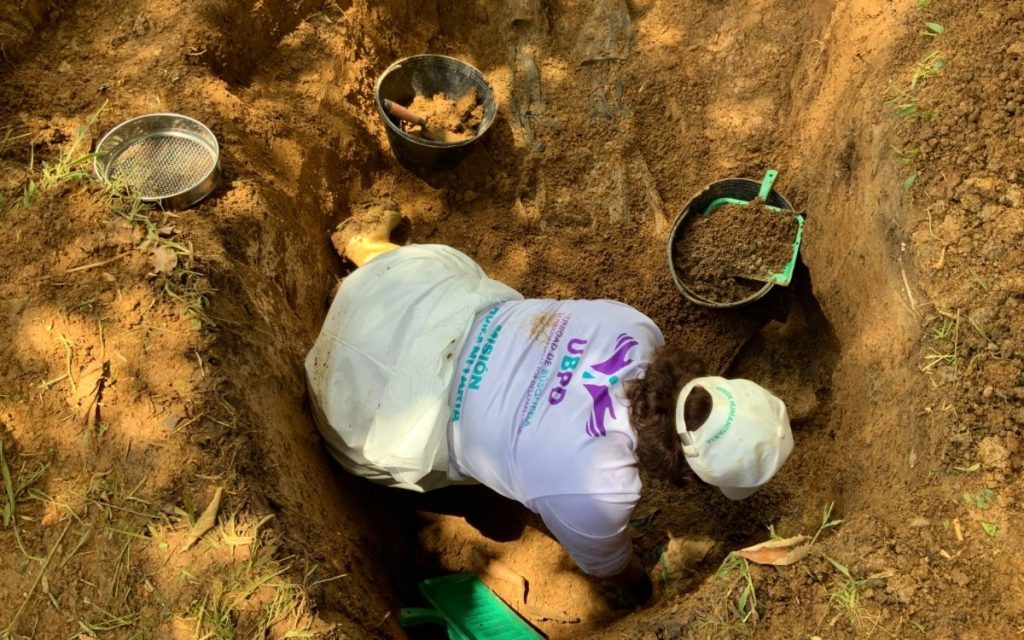 Plano cenital (desde arriba) que muestra a una profesional de la UBPD inclinada dentro de una fosa o excavación en el terreno. Viste una camiseta blanca con el logotipo de la UBPD en la espalda, una gorra clara y guantes de protección. A su alrededor, sobre la tierra, hay utensilios de trabajo forense y arqueológico, como un cernidor o tamiz metálico redondos, baldes y palustres para remover tierra con precisión.