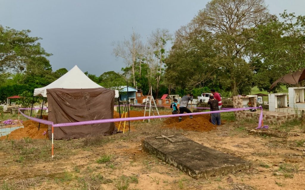 Toma abierta de un cementerio rural al aire libre. En la parte central, un área de terreno está acordonada con cinta sintética rosada. Dentro del perímetro, varios trabajadores realizan excavaciones manuales con palas junto a montículos de tierra extraída. Hay instalada una carpa blanca de campamento con lonas laterales para proteger la zona de trabajo, además de una escalera de aluminio y herramientas. En el primer plano se aprecian tumbas y placas de cemento sobre el suelo rodeadas de vegetación y árboles altos.