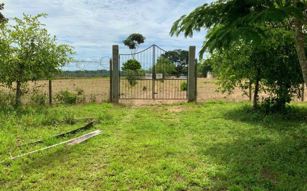 Portón metálico de dos hojas sostenido por pilares de concreto que da acceso a un terreno plano o cementerio rural rodeado por un cerco de malla de alambre. El área exterior está cubierta de pasto verde y vegetación, bajo un cielo parcialmente nublado.