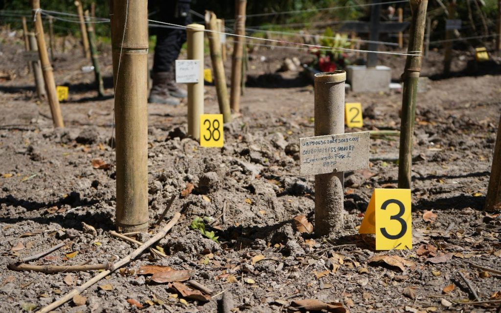 Detalle a ras de suelo en el que destacan marcadores de evidencia amarillos numerados (3 y 38) junto a estacas de bambú delimitadas con cuerdas. En el centro, una tablilla clavada en la tierra registra un código técnico de intervención de la UBPD.