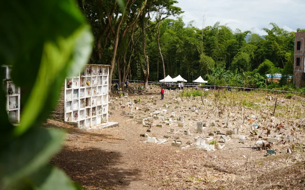 Plano general que muestra a la izquierda una estructura vertical de bóvedas funerarias blancas. El terreno abierto, con cruces y tumbas a ras de suelo, conduce hacia el fondo donde se aprecian carpas blancas de intervención forense delimitadas con cintas de seguridad, rodeadas por un denso bosque de árboles y guaduas.