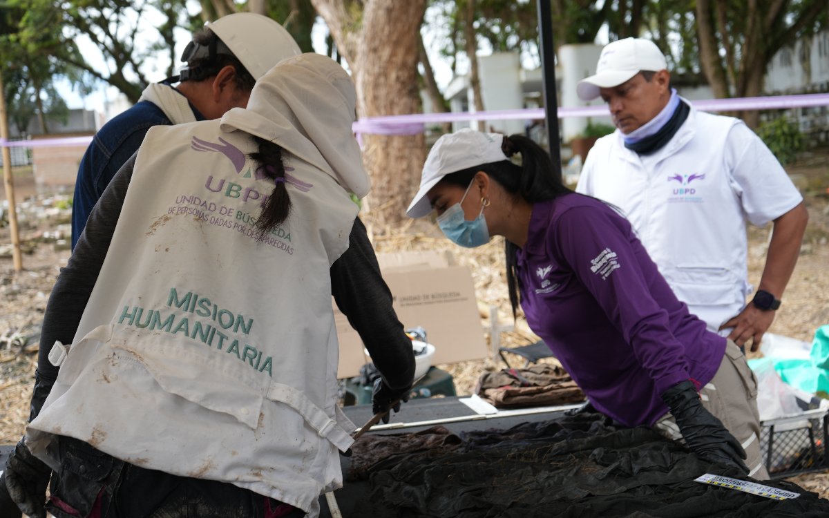 Tres profesionales de la Unidad de Búsqueda de Personas dadas por Desaparecidas (UBPD) trabajan en campo. Una mujer con gorra y tapabocas revisa minuciosamente prendas de vestir oscuras extendidas sobre una mesa, acompañada por un colega con chaleco blanco institucional y otro observando de pie.