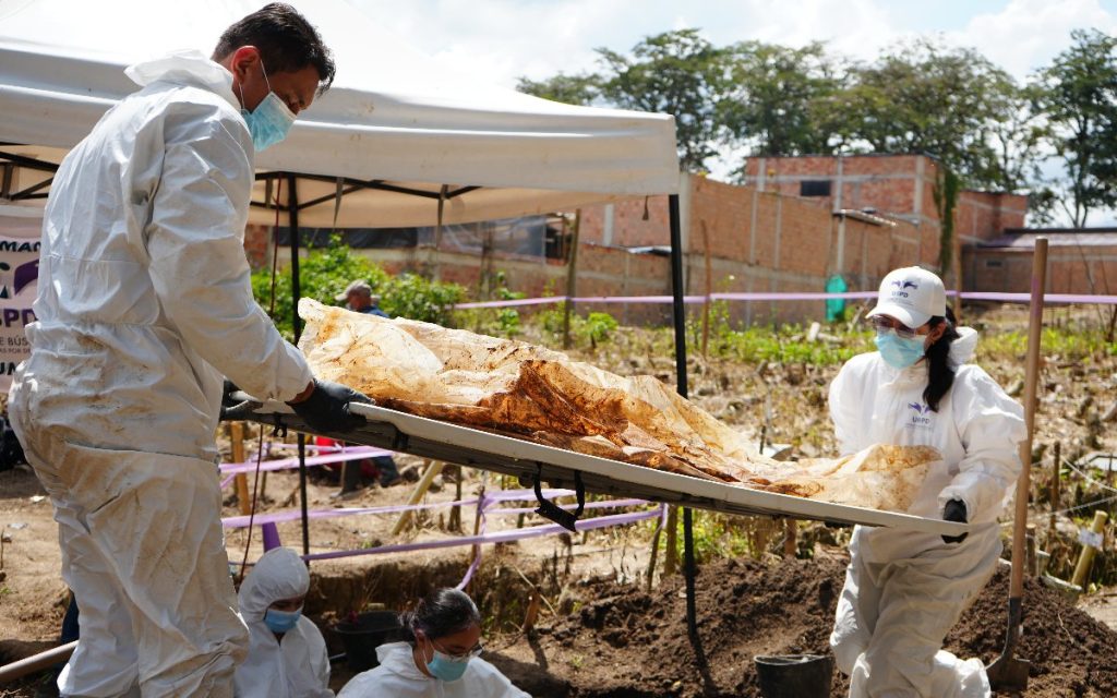 Dos funcionarios con trajes biológicos completos blancos, guantes y tapabocas trasladan una bandeja o camilla metálica con evidencias envueltas en plástico y tierra. Al fondo se observa una carpa de trabajo y cinta perimetral morada en el terreno del cementerio.
