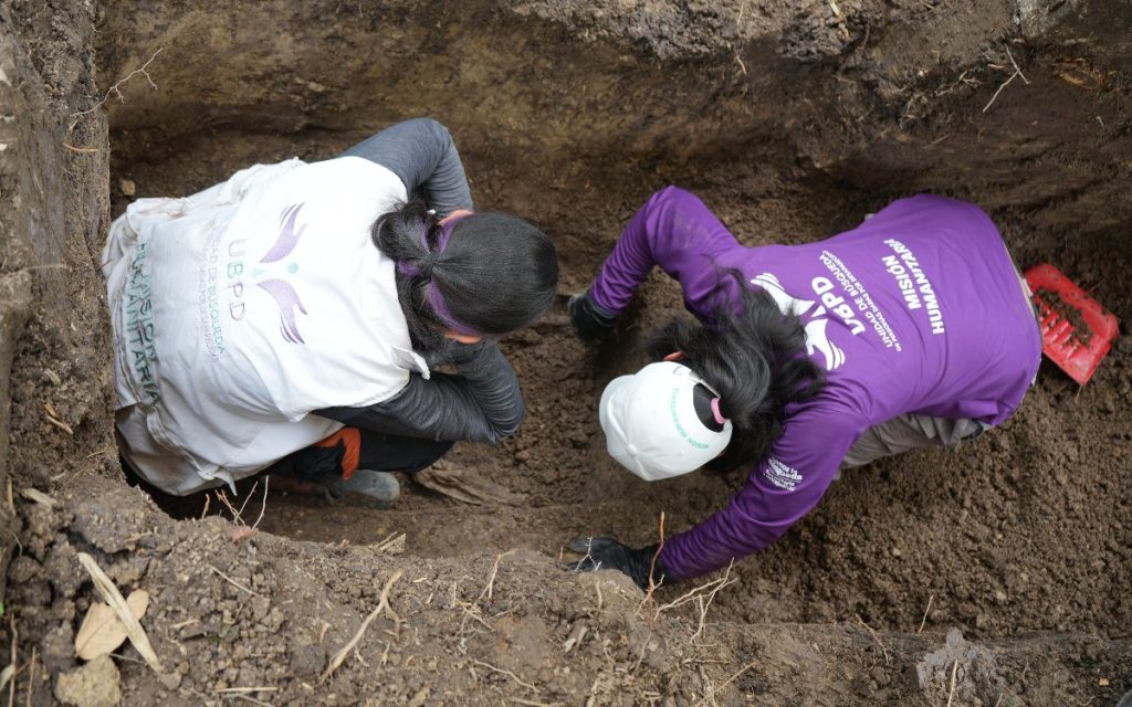 Toma en picado donde dos funcionarias de la UBPD (una con chaleco blanco y otra con camisa morada institucional) remueven tierra con guantes de protección en el fondo de una fosa excavada en el suelo.