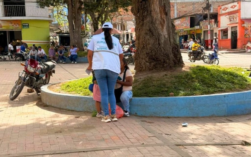 Una funcionaria vista de espaldas, con chaleco blanco identificativo de la Unidad de Búsqueda de Personas dadas por Desaparecidas (UBPD) con el texto "Misión Humanitaria - Somos la búsqueda", dialoga con una persona en un parque al aire libre. La escena se desarrolla junto al tronco de un árbol grande sobre una jardinera de borde azul. Al fondo se observan motocicletas, otras personas sentadas y locales comerciales.