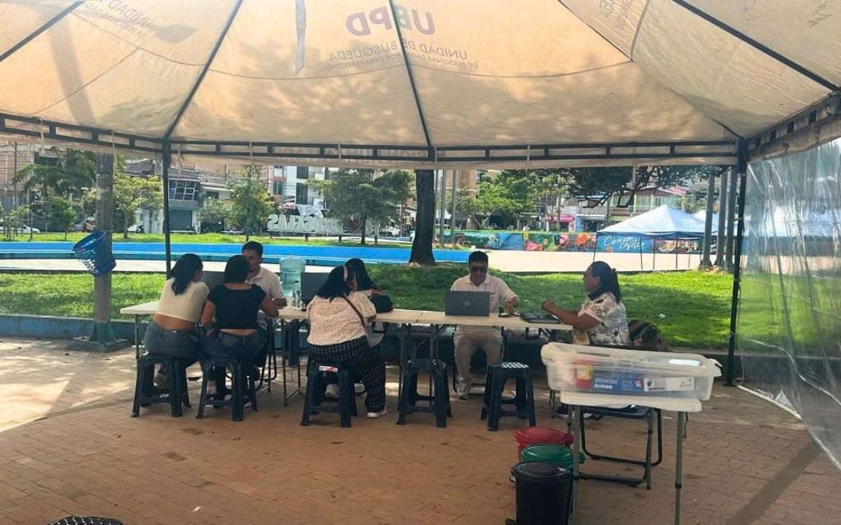 Interior de una carpa institucional blanca ubicada en un parque arbolado. Varios profesionales de la Unidad de Búsqueda atienden a ciudadanos sentados en mesas plegables con computadores portátiles. Sobre una mesa auxiliar se aprecia una caja de almacenamiento con insumos institucionales.