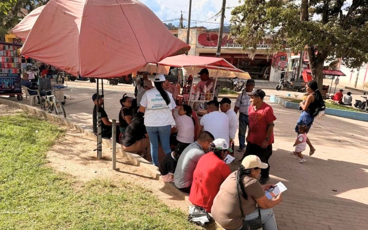 Punto de atención al aire libre protegido por grandes parasoles o sombrillas rojas. Integrantes de la Unidad de Búsqueda con chalecos institucionales conversan con miembros de la comunidad que se encuentran sentados en el sardinel y en sillas, algunos leyendo folletos informativos.