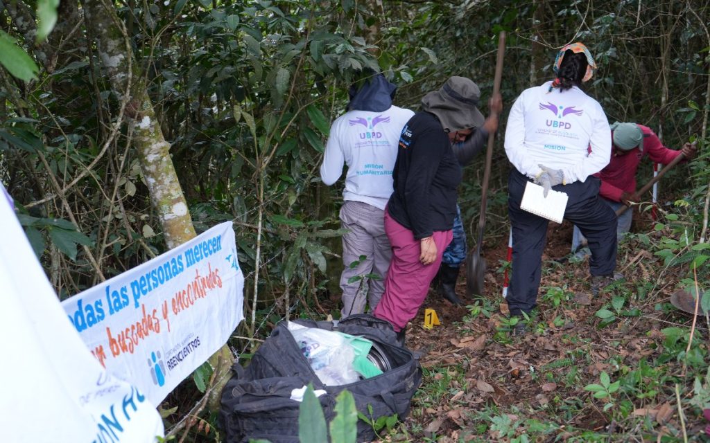 En una zona boscosa rodeada de abundante vegetación y árboles, cuatro integrantes de la Unidad de Búsqueda de Personas dadas por Desaparecidas (UBPD) y personal de apoyo realizan labores de excavación en el terreno. Tres de ellos vestidas con camisetas blancas con el logo de la UBPD y la leyenda "Misión Humanitaria". A la izquierda se observa una pancarta blanca con el mensaje "Todas las personas merecen ser buscadas y encontradas - Corporación Humanitaria Reencuentros". Junto a la zona de excavación hay una maleta negra de herramientas y un señalizador amarillo con el número 1.