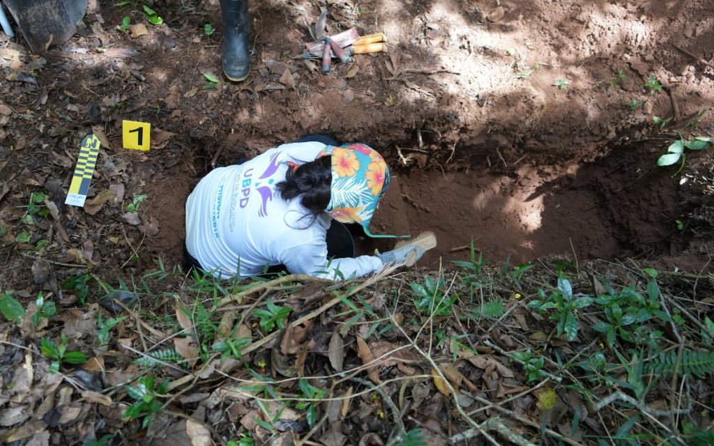 Toma aérea directa hacia un punto de excavación forense. Una integrante de la UBPD, que viste camiseta blanca de la institución y pañoleta de flores en la cabeza, se encuentra agachada al borde de una trinchera o fosa de tierra utilizando un cepillo para retirar sedimentos de manera cuidadosa. Fuera de la fosa, sobre la hojarasca del suelo, se aprecia una baliza con el número 1, una escala métrica amarilla y negra, y herramientas de mano depositadas sobre la tierra.