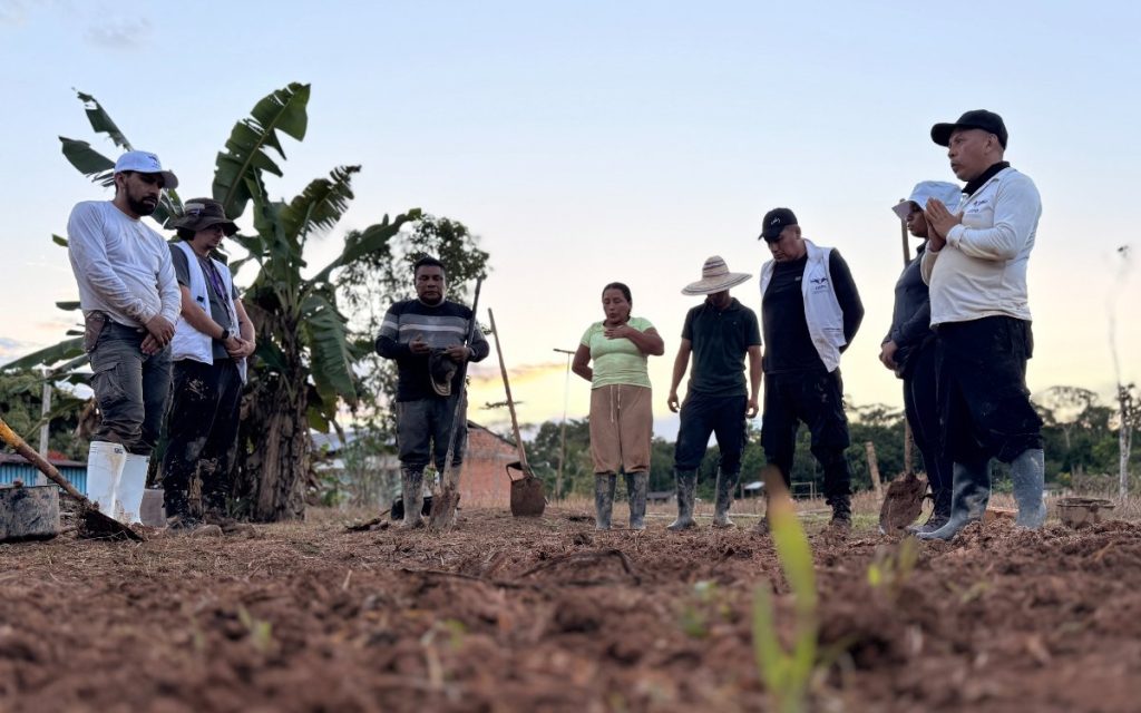 Un grupo de nueve personas se encuentra de pie en un semicírculo en un terreno de tierra arada al aire libre, con vegetación y árboles de plátano al fondo bajo un cielo claro al atardecer. Varias de las personas visten botas pantaneras y algunas llevan chalecos o gorras de trabajo, manteniendo posturas solemnes y de respeto, con las manos juntas o en el pecho como en un momento de oración o conmemoración. En los extremos y en el suelo se observan palas y herramientas de excavación clavadas en la tierra. Un hombre a la derecha, con gorra y camiseta blanca, habla al grupo con las manos juntas.