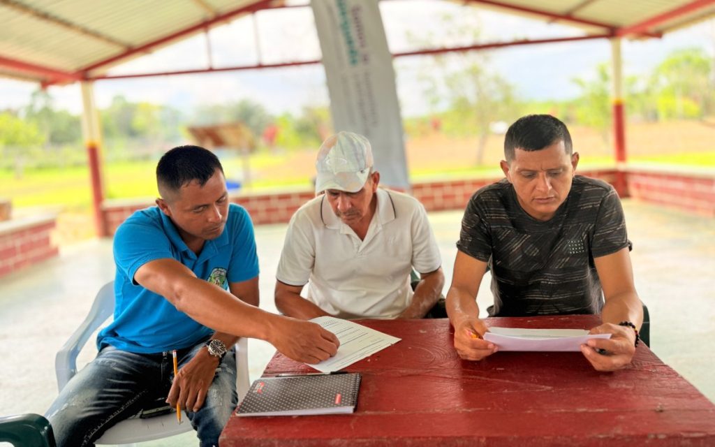 Tres hombres sentados alrededor de una mesa rústica de color rojo bajo una estructura abierta con techo de zinc en una zona rural. A la izquierda, un hombre con camiseta azul señala con un lápiz un documento en papel sobre la mesa mientras el hombre del centro, con gorra clara y camiseta blanca, lo observa con atención. A la derecha, un tercer hombre con camiseta oscura y de espaldas parciales lee concentrado otro documento que sostiene con ambas manos. Sobre la mesa se aprecian un cuaderno de apuntes y hojas sueltas, con un entorno natural y edificaciones de fondo de manera difuminada.