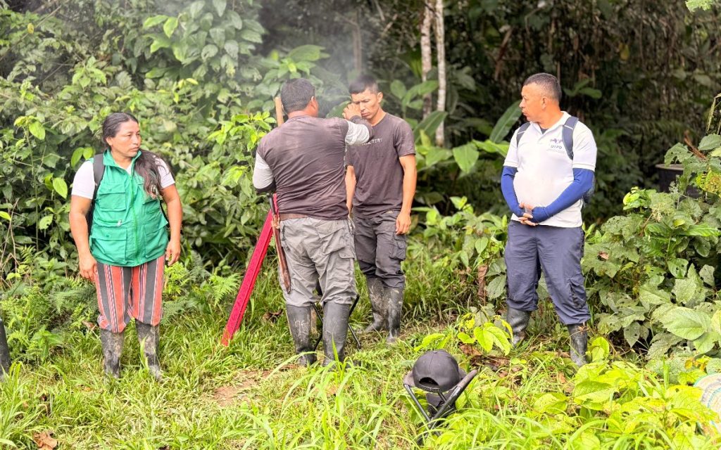 Cuatro personas de pie en una zona de densa vegetación y maleza verde. A la izquierda, una mujer con chaleco verde de brigada y pantalón a rayas observa hacia la derecha. En el centro, un hombre de espaldas con camiseta oscura y pantalón gris con botas de caucho atiende a otro hombre con camiseta negra que se toca el rostro, cerca de un trípode de medición rojo. A la derecha, un hombre con camiseta blanca y mangas azules que lleva el logo "UBPD" en el pecho y un morral los observa con las manos entrelazadas. En el suelo se ve vegetación abundante y una pequeña silla plegable con una gorra encima.