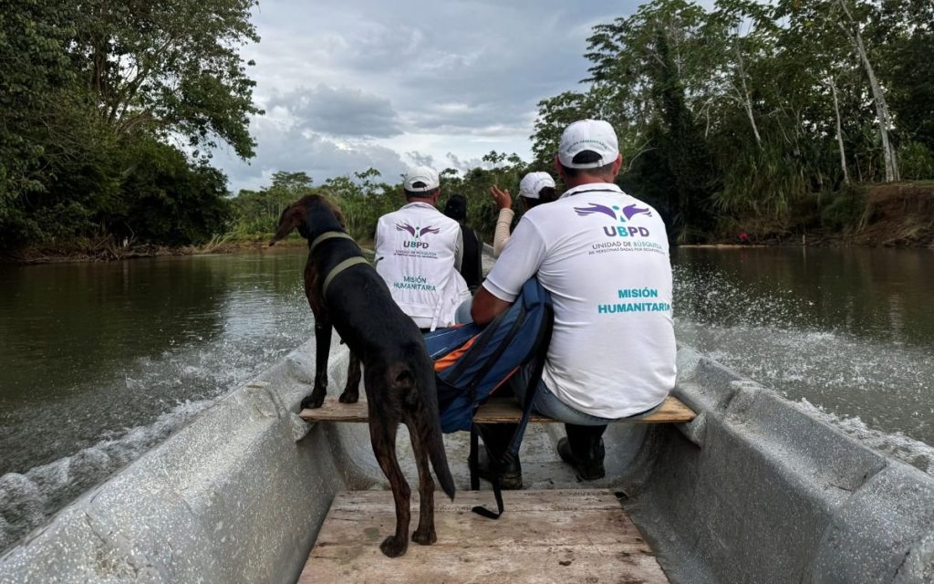 Fotografía desde la parte trasera de una embarcación de madera sobre un río navegable. En primer plano, de espaldas, hay un perro negro de pie observando hacia adelante. A su lado viajan dos personas vistiendo prendas con el logo de la UBPD y el texto "Misión Humanitaria". De fondo se aprecian árboles y vegetación densa a las orillas del río bajo un cielo nublado.