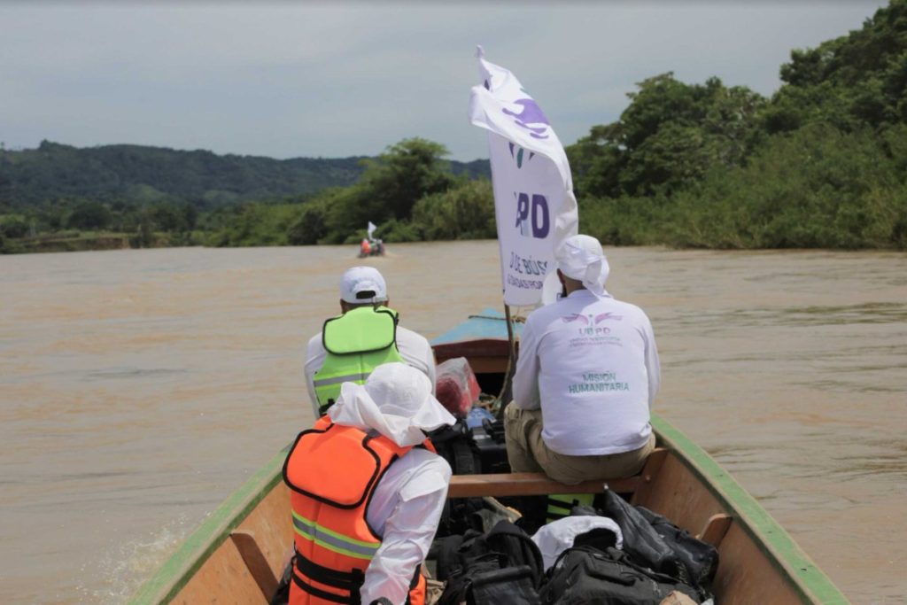 Vista trasera desde el interior de una lancha de madera en movimiento sobre un río turbio de color marrón. Tres personas vistiendo ropa blanca de manga larga y chalecos salvavidas fluorescentes viajan a bordo. Una bandera blanca con el logo de la UBPD ondea cerca de la proa. Al fondo se observa otra embarcación navegando, colinas verdes y vegetación en las márgenes del río.