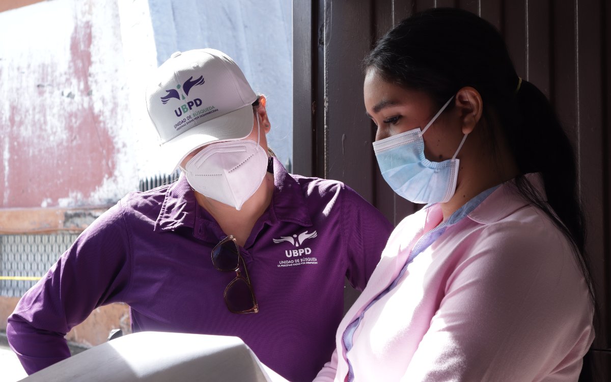 Plano medio de dos funcionarias trabajando en campo con mascarillas protectoras. A la izquierda, una integrante de la Unidad de Búsqueda de Personas dadas por Desaparecidas (UBPD) con uniforme institucional morado y gorra blanca observa atentamente unas hojas junto a una compañera.