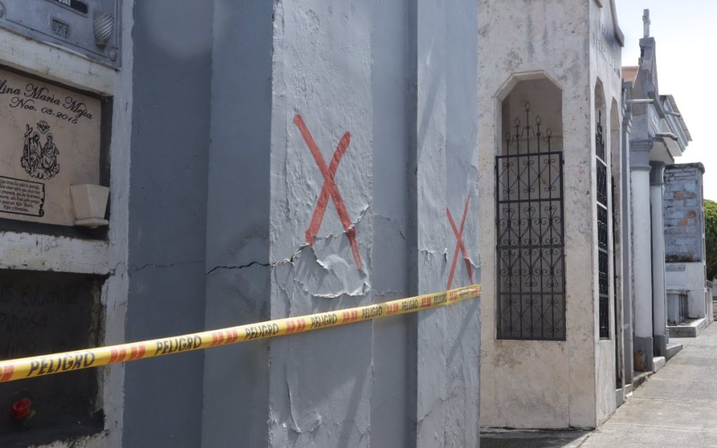 Espacio de oficina donde se observan dos personas en labores de archivo y custodia documental. En primer plano, de espaldas, se distingue el logo institucional de la UBPD en una camisa morada; al fondo, un colaborador con tapabocas y guantes manipula carpetas junto a cajas rotuladas sobre un escritorio de trabajo.