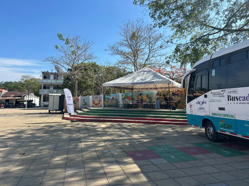 Fotografía en un día despejado de una plaza o parque público. En el costado derecho de la imagen aparece la parte delantera de un autobús blanco y azul con los logotipos de la Unidad de Búsqueda de Personas dadas por Desaparecidas (UBPD) y el texto «La ruta Buscadora - Unidad Móvil». Al fondo, sobre una tarima con escalones decorados con franjas de colores, se encuentra instalada una carpa institucional blanca con la inscripción «Misión Humanitaria». Detrás de la carpa se observan murales artísticos coloridos y árboles. El piso de la plaza está pavimentado con losas de piedra y en el suelo en primer plano hay dibujado un juego infantil.