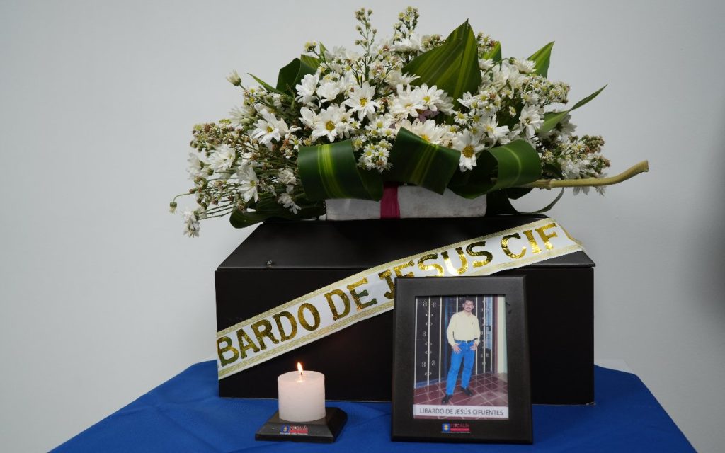 Cofre fúnebre con cinta que dice «Libardo de Jesús Cifuentes», cubierto por un arreglo de flores blancas, acompañado al frente por una vela encendida y un portarretratos con la fotografía de un hombre joven en camisa clara y pantalón azul.   