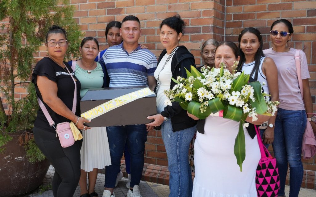 Plano medio de familiares rodeando y sosteniendo el cofre fúnebre con la cinta de Libardo de Jesús Cifuentes y un gran ramo de flores blancas frente a un muro de ladrillo.