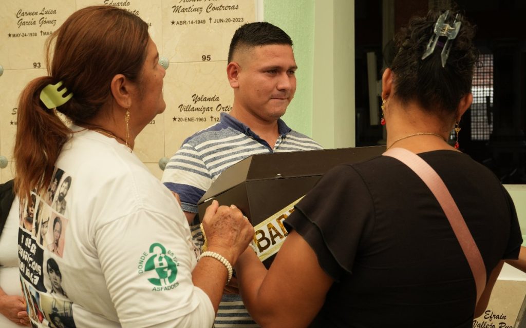 Un hombre joven y dos familiares sostienen el cofre fúnebre en el área de bóvedas del cementerio; una de las mujeres lleva una camiseta con el logo de ASFADDES.