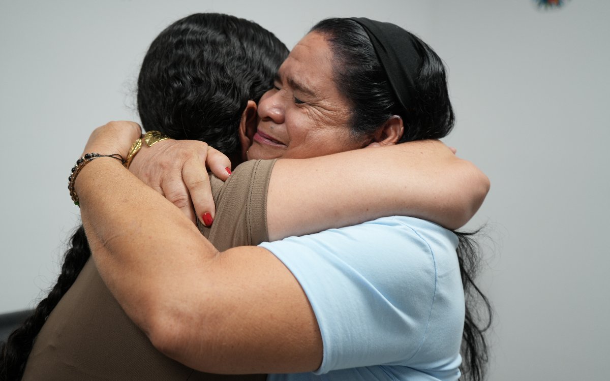 Fotografía en plano detalle de un abrazo entre dos mujeres. Una mujer de cabello largo y negro con balaca abraza fuertemente a otra, recostando su rostro con expresión emotiva y los ojos cerrados.
