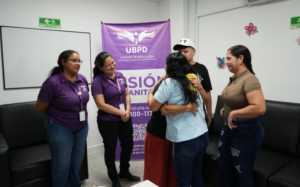 En una oficina con el pendón institucional de la UBPD al fondo, un grupo de familiares se reúne en un abrazo emotivo. A la izquierda, dos funcionarias con camisetas moradas del logo de la UBPD presencian la escena con expresión de alegría y empatía.