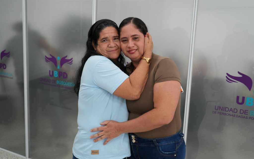 Retrato de medio cuerpo de dos mujeres sonrientes abrazadas fraternalmente frente a una pared divisoria de vidrio que lleva el logo de la Unidad de Búsqueda de Personas dadas por Desaparecidas (UBPD). Una viste camiseta azul clara con balaca negra y la otra viste camiseta café.
