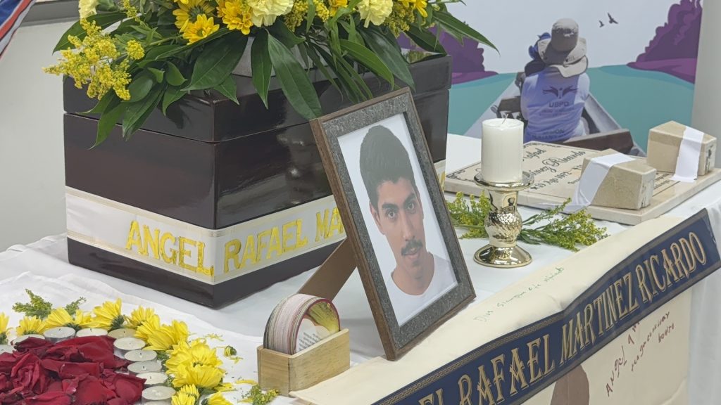Plano corto que muestra los elementos en memoria de Ángel Rafael Martínez Ricardo: un cofre funerario de madera oscura con su nombre grabado en letras doradas, un retrato enmarcado, pétalos de rosas rojas y arreglos de flores amarillas rodeados por velas.