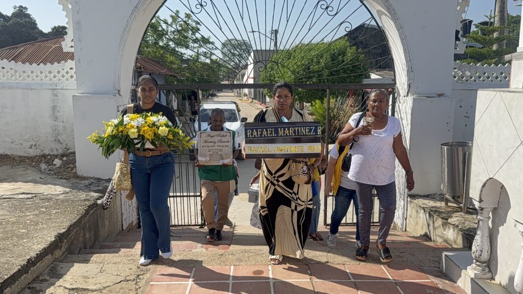 Familiares ingresando a través de un portón de reja de hierro blanco al cementerio. Una mujer encabeza el recorrido con un ramo floral amarillo y otra persona tras ella porta la urna ceremonial cubierta con una tela con el nombre de Ángel Rafael Martínez Ricardo.