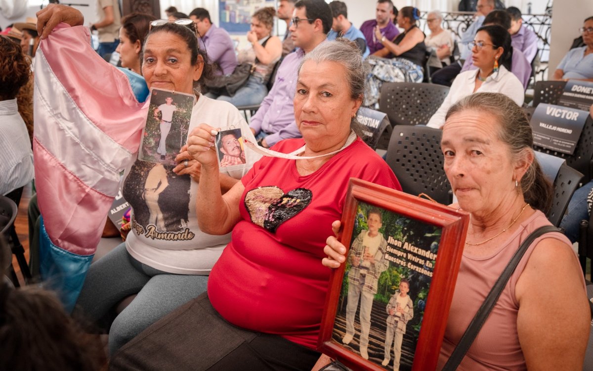 Plano medio de tres mujeres asistentes a un evento público. La mujer de la izquierda sostiene una bandera trans de colores rosa, blanco y azul, así como una fotografía impresa de un joven. La mujer del centro viste camiseta roja y sostiene una pequeña foto. La mujer de la derecha porta un cuadro enmarcado en madera con la fotografía de un niño e inscripciones conmemorativas. Al fondo se observa a otros asistentes en el auditorio.