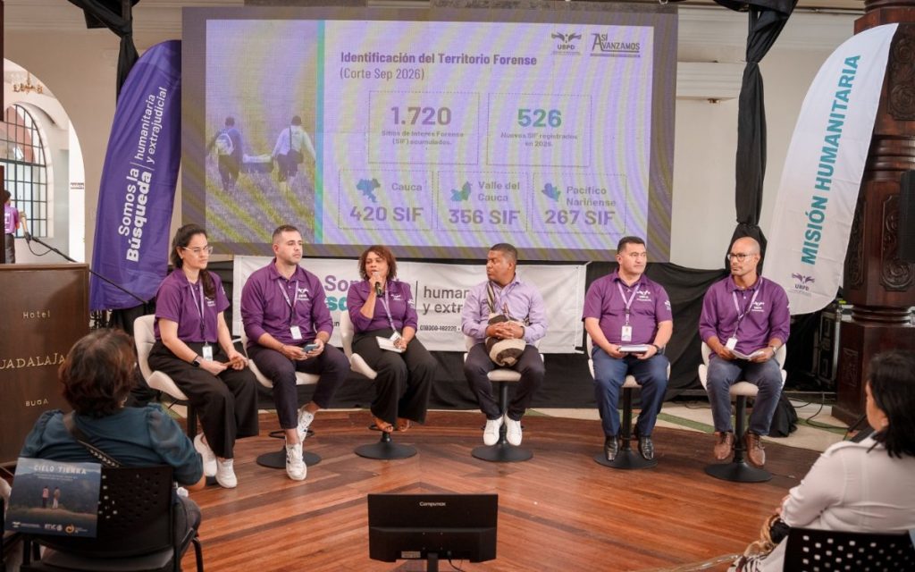 Vista del escenario con cuatro panelistas sentados. A la izquierda, un hombre habla por un micrófono sosteniendo documentos; a su lado está una mujer con vestido verde, seguida de dos hombres con camisas lilas. Al fondo, la pantalla exhibe el título "Más capacidad forense para identificar y entregar respuestas", detallando seis nuevos CIAFI articulados, 417 procedimientos, 1.047 cuerpos abordados y 107 identificaciones logradas. En primer plano se ven las cabezas de asistentes, entre ellos personas con sombreros tradicionales.   