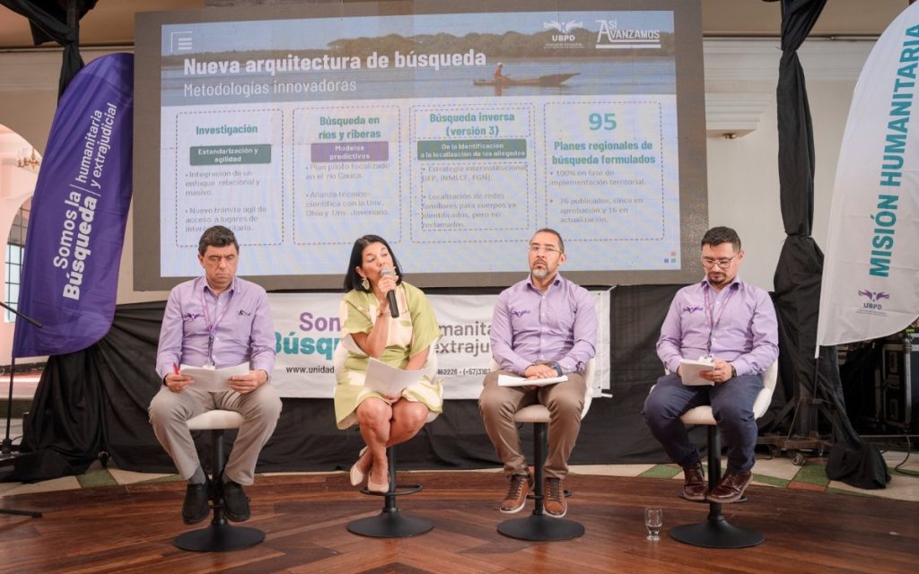 Fotografía en plano frontal de cuatro panelistas sentados en taburetes. Una mujer vestida de verde habla por el micrófono rodeada por tres hombres con camisas de la institución. La pantalla gigante a su espalda muestra un esquema informativo titulado "Nueva arquitectura de búsqueda - Metodologías innovadoras", dividido en secciones: Investigación, Búsqueda en ríos y riberas, Búsqueda inversa (versión 3) y 95 Planes regionales de búsqueda formulados.