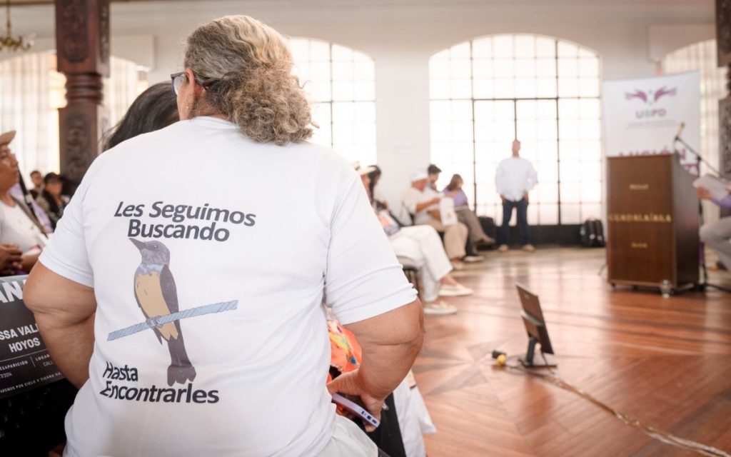Fotografía panorámica del evento de rendición de cuentas de la UBPD. En primer plano se observa al público asistente de espaldas, compuesto por hombres y mujeres de diversas edades. Al fondo, sobre un escenario de madera flanqueado por columnas de madera labrada y pendones institucionales, se encuentran cuatro integrantes del panel y un moderador en el atril a la izquierda. En la pantalla central se lee el logo de la UBPD con el lema "Así Avanzamos - Audiencia pública rendición de cuentas".